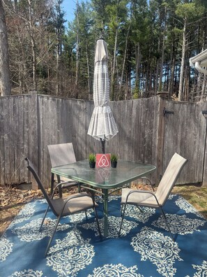 Een glazen tafel met twee stoelen en een parasol, geplaatst op een tapijt met een patroon, met een houten omheining en bomen op de achtergrond.