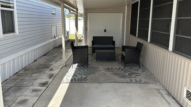 Ein überdachter Patio-Bereich mit einer Couch und zwei Stühlen, einem Betonboden und Blick auf ein Haus mit Garagentor.