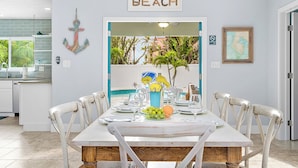 Uma área de refeições com mesa posta para seis pessoas, uma âncora decorativa na parede, e uma placa acima da porta que diz "BEACH".