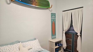 A bedroom with a bed, a starfish pillow, a surfboard decoration, a surf shack sign, and a window with curtains. - Cocoa Beach Condo - SeaBreeze (Cocoa Beach)