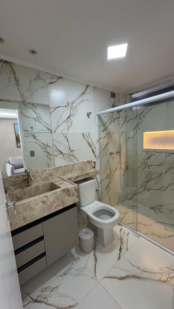 A modern bathroom with a marble backsplash, a glass shower enclosure, and a toilet. - Flat luxo enseada (Maceio)