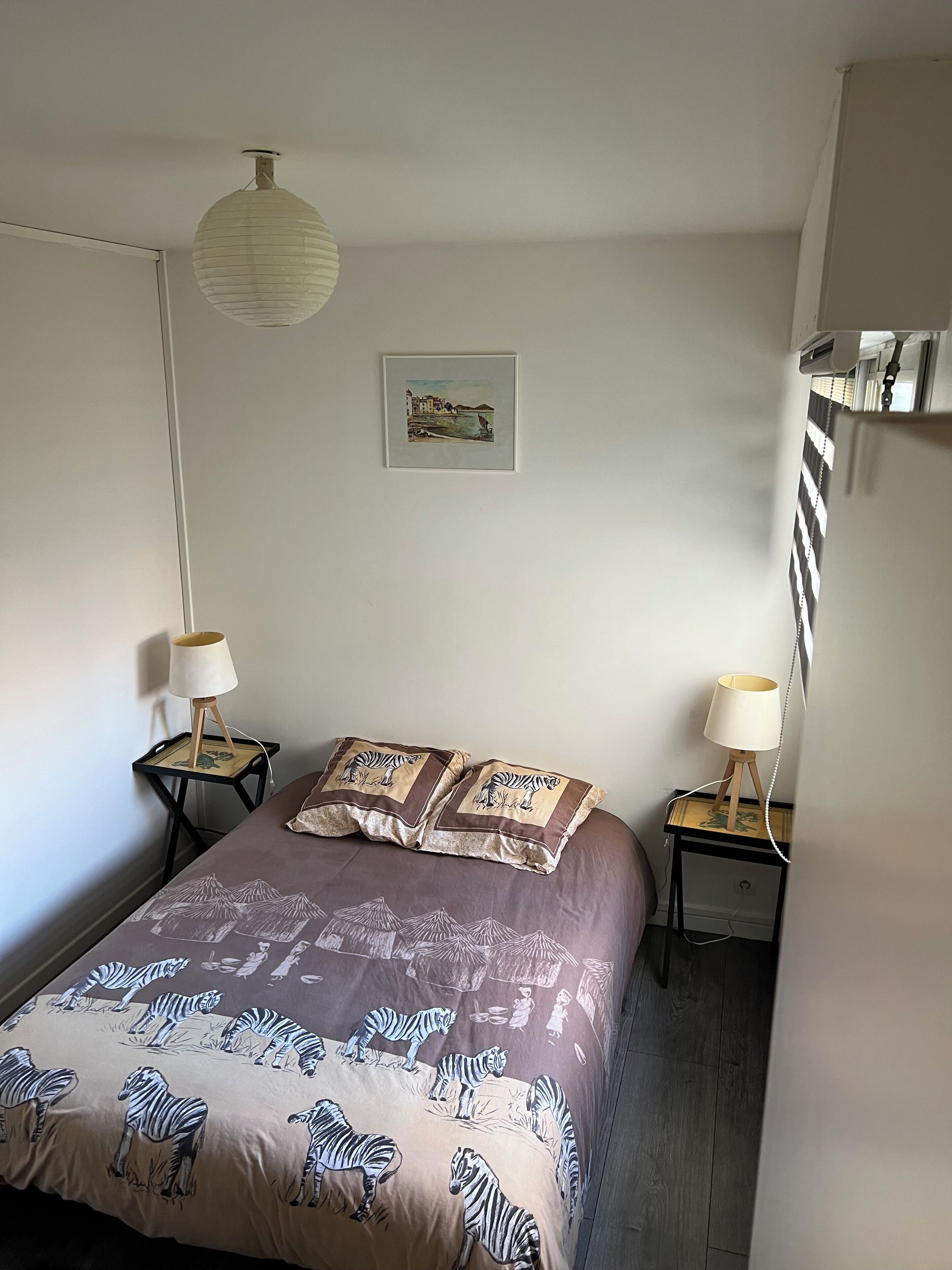 A bedroom with a bed, two bedside tables with lamps, a hanging light fixture, a framed picture on the wall, and a wooden floor.