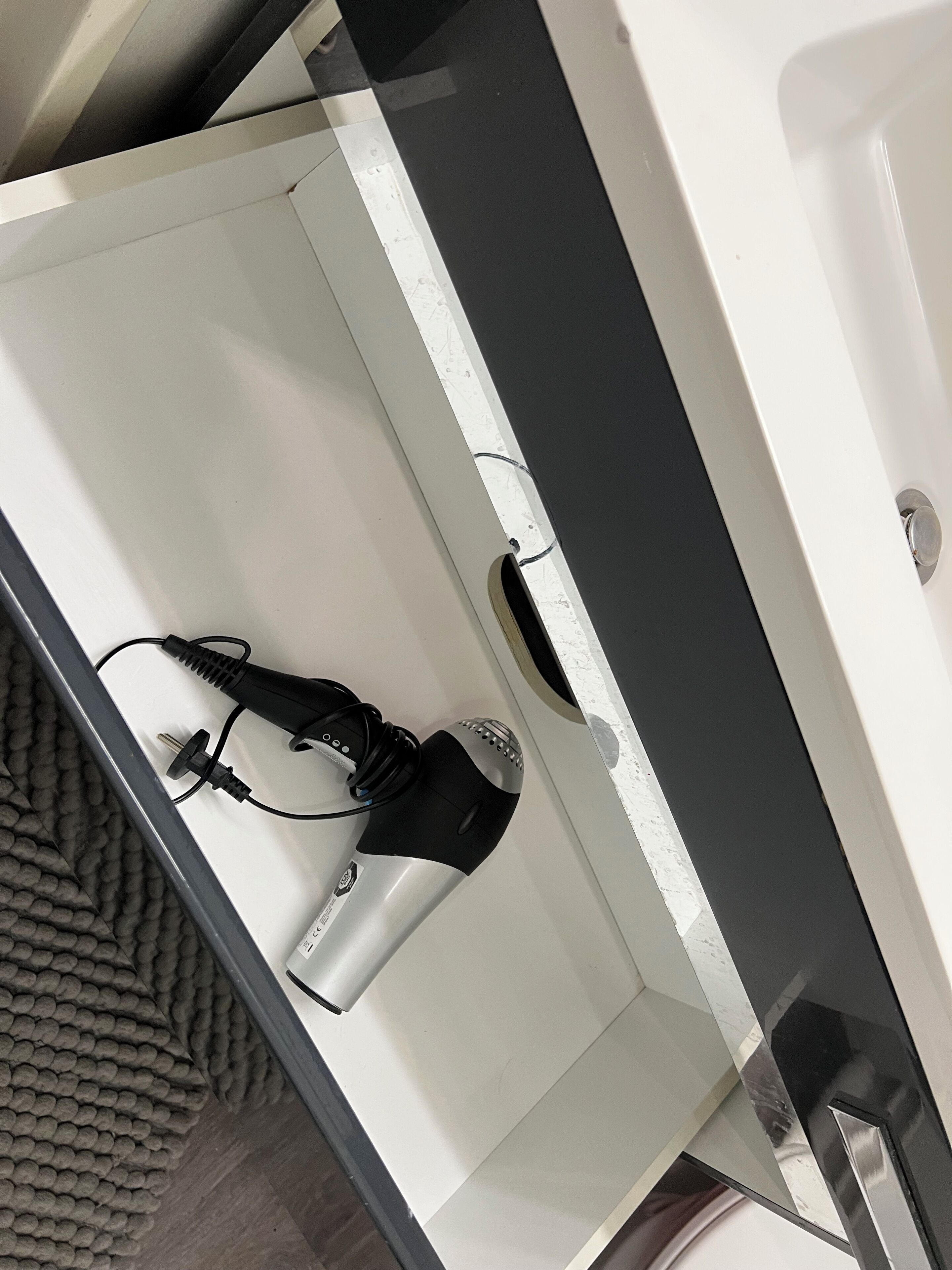 Una habitación blanca con un secador negro sobre un estante blanco, un secador negro y plateado en el suelo, y una alfombra gris con textura.
