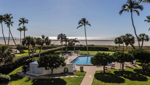 Zona de piscina con vista a la playa, rodeada de palmeras y un césped cuidado.