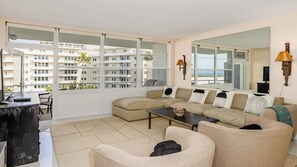 A living room with a sofa, armchairs, a coffee table, and a TV. There are large windows with a view of buildings and palm trees.
