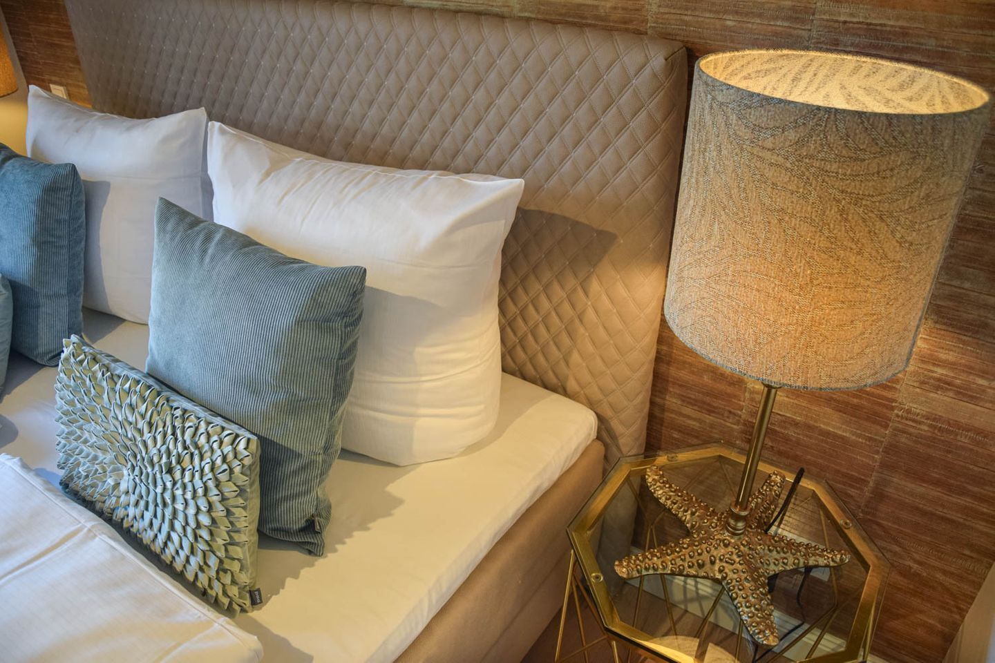 Una cama con cabecera acolchada, varios cojines, una lámpara con forma de estrella de mar y una mesita de cristal con un objeto decorativo.