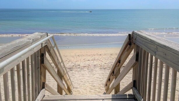 Una pasarela de madera bordeando una playa arenosa, con vista despejada al océano.