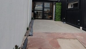 Un camino pavimentado que conduce a un edificio de diseño moderno, con un vestíbulo cubierto y un pequeño jardín con flores rosadas.