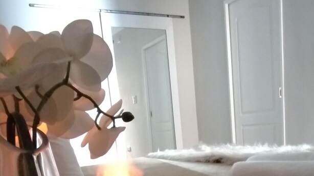 Una habitación con una mesita de noche, un jarrón con flores blancas, un calentador eléctrico encendido, una cama con ropa blanca y una puerta blanca al fondo.