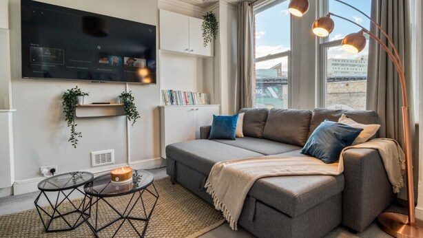 A modern living room with a grey sectional sofa, a flat-screen TV mounted on the wall, a small round coffee table, and a floor lamp. - Chic 1Bed w/Patio Battersea (Greater London)