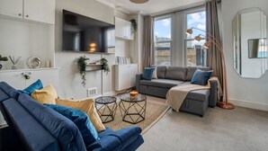 A modern living room with a grey sofa, a blue sofa, a TV mounted on the wall, and a large window with curtains. - Chic 1Bed w/Patio Battersea (Greater London)