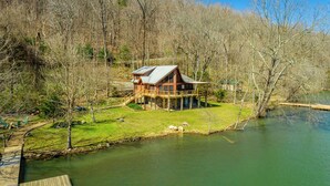 Een chalet met een stenen fundering, een houten terras en een groot raam met uitzicht op een rivier.