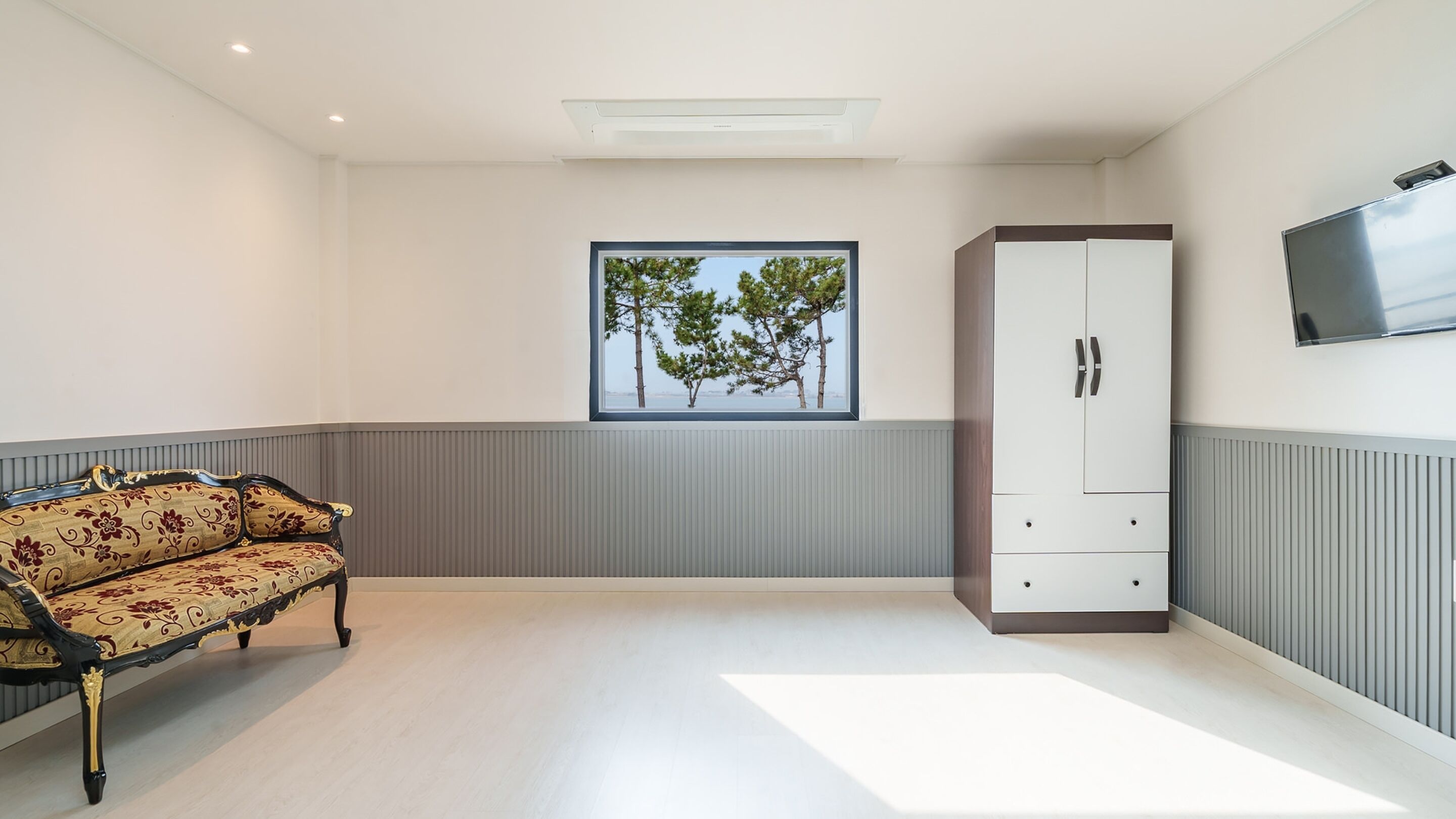 Ein Zimmer mit einer Couch, einem Schrank, einem Fenster mit Blick auf Bäume und einem an der Wand montierten Flachbildfernseher.
