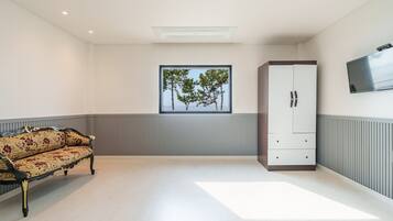 A room with a sofa, a cabinet, a window with a view of trees, and a flat-screen TV mounted on the wall.
