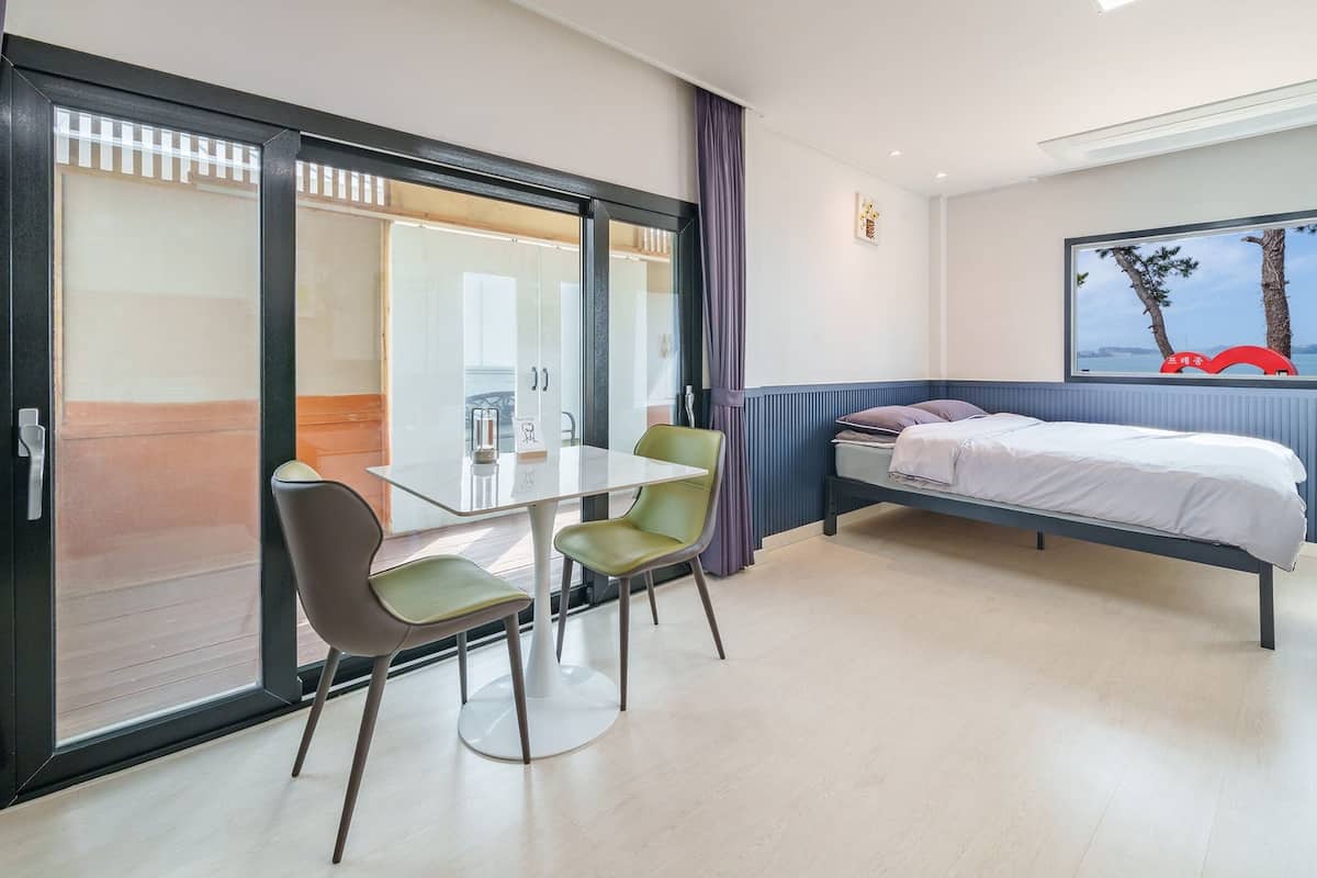 A hotel room with a bed, a table with two chairs, a sliding glass door, and a painting of a tree and a red chair on the wall.