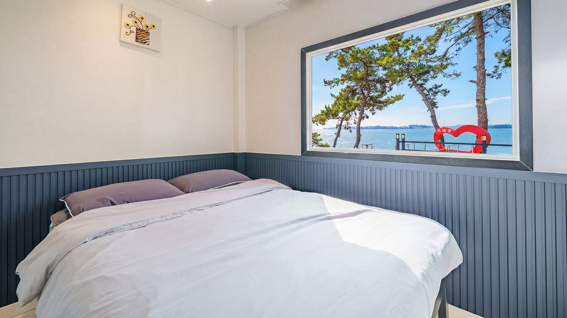 A bedroom with a bed, a window with a view of trees and water, and a wall-mounted picture.