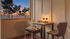 A balcony with a table set for two, a grill with food, and a view of trees and water at sunset. - Mu-an Prejong Dokchae Pension (Muan)