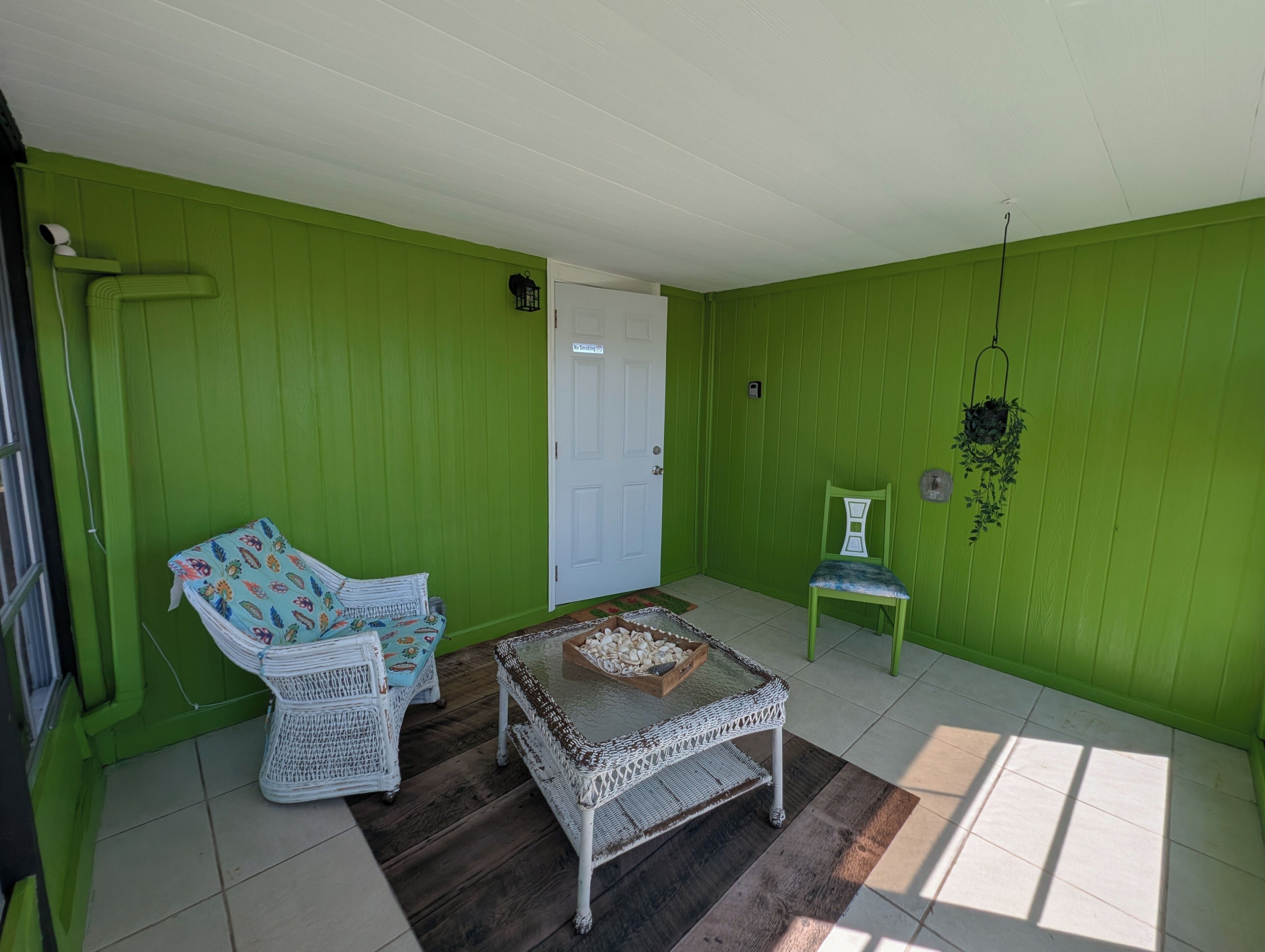 A room with green walls, a white door, a wicker chair with a floral cushion, a glass-top table with a decorative bowl, and a hanging plant.
