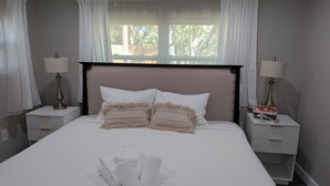 Una cama bien hecha con ropa blanca, dos mesitas de noche con lámparas y una ventana con cortinas.
