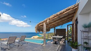 Une terrasse dotée d’un toit de chaume, une piscine, des chaises longues et une table avec des chaises.