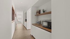 Un couloir étroit, doté d’un plancher en bois, mène à une cuisine avec des meubles blancs et un four à micro-ondes posé sur le plan de travail.