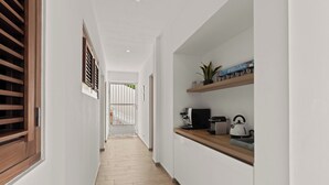Un couloir étroit doté d’un plancher en bois, de murs blancs et d’un coin cuisine avec un plan de travail, une bouilloire et des étagères.