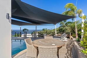 Een zwembadterras met een ronde houten tafel en rieten stoelen, met uitzicht op het zwembad en een schilderachtig uitzicht over de oceaan.