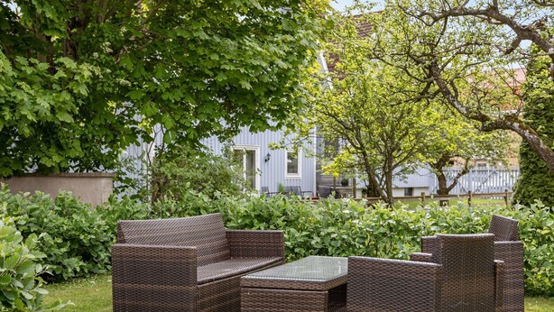 En utomhusplats med sittmöbler i rotting, ett glasbord och frodig grönska och träd runt omkring.