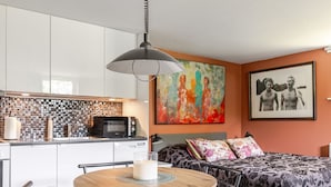 Ett kompakt bostadsutrymme med ett litet kök, en matplats med ett runt bord och stolar, en säng med blommigt sänglinne och en färgstark, abstrakt tavla på väggen.