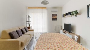 Un dormitorio moderno con sofá, cama con colcha estampada, televisor sobre un mueble bajo y ventana con cortinas ligeras.
