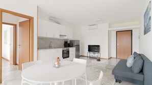 A modern kitchen with white cabinets, a dining table with chairs, a gray sofa, and a TV on a stand.