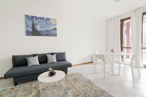 A modern living room with a grey sofa, a round coffee table, a white dining table with chairs, and a painting on the wall.
