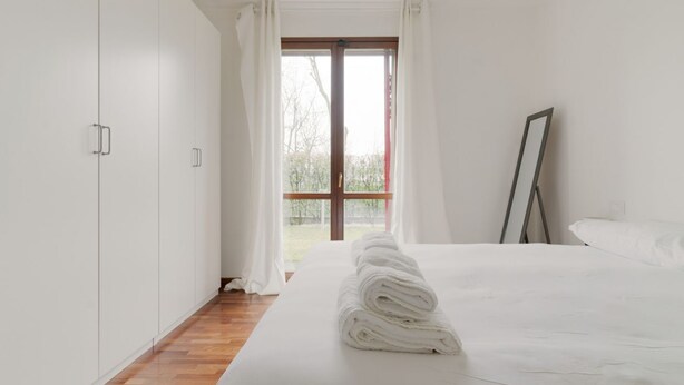 A bedroom with a large bed, white curtains, a window, and a wooden floor.