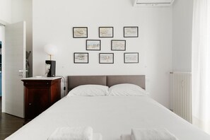 Habitación de hotel con una cama grande, una mesita de noche con lámpara, un radiador y cuadros en la pared.