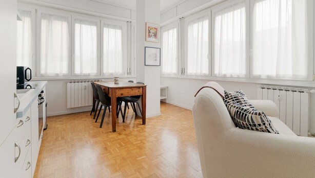 A bright room with a dining table, chairs, a sofa, and large windows with curtains. - GuestHost - Cozy 78 sqm apartment, able to accommodate up to 4 people, located on the first floor of a building with lift.The property is located 650m from the Famagosta stop of the green metro line M2, which allows you to reach Piazza Duomo, Central Sta (Milano)