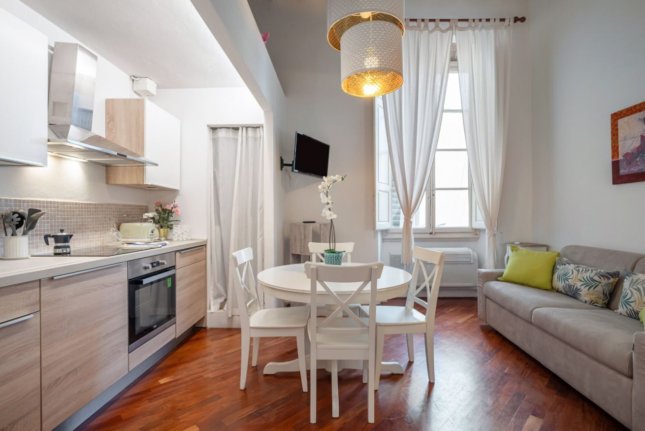 A modern kitchen with white cabinetry, a dining area with white chairs, a sofa with patterned cushions, and a large window with curtains.