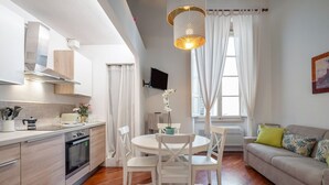 Eine moderne Küche mit weißen Schränken, ein Essbereich mit weißen Stühlen, eine Couch mit gemusterten Kissen und ein großes Fenster mit Vorhängen.