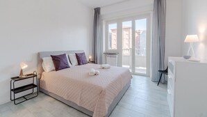 A modern hotel room with a bed, a nightstand, a desk, and a view of buildings through the window.