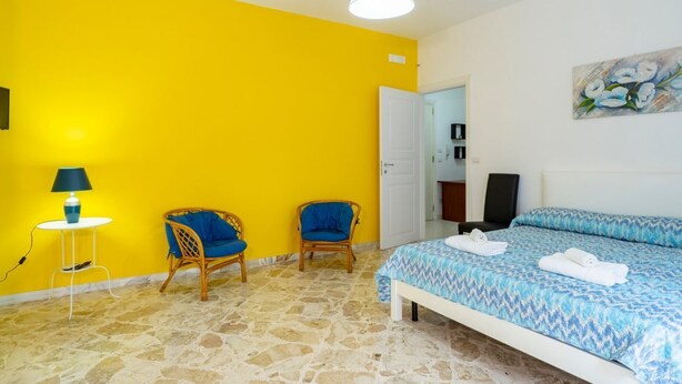 A room with a yellow wall, a bed with a patterned blue bedspread, two wicker chairs, a white bedside table with a lamp, and a painting on the wall.