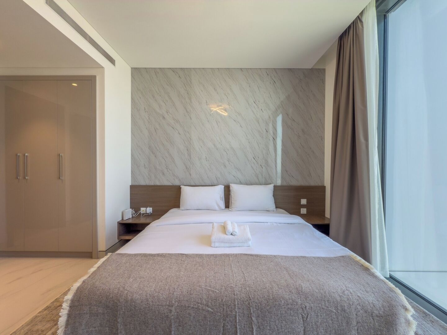 Une chambre d’hôtel moderne, dotée d’un grand lit, d’une table de chevet, d’une armoire et offrant une vue sur la ville par la fenêtre.