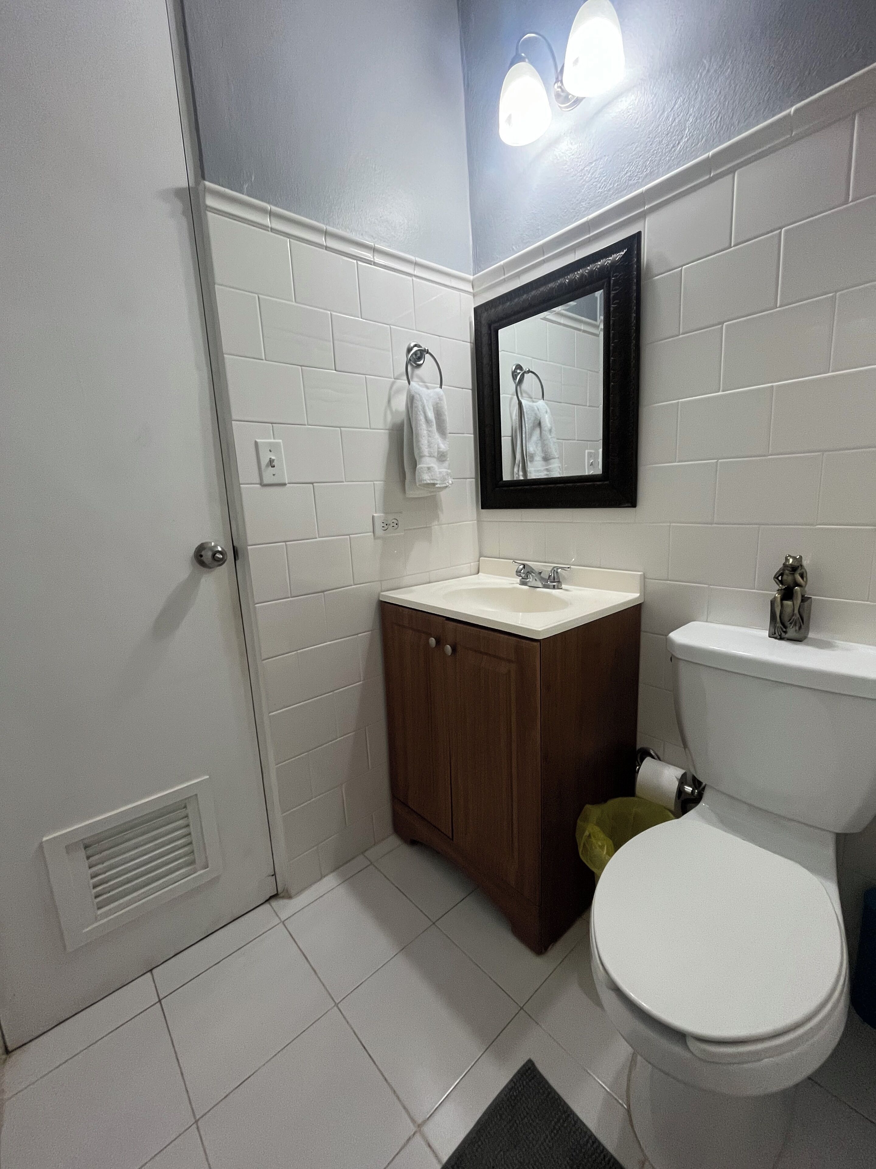 Ein Badezimmer mit weiß gefliesten Wänden, einem Holzschrank, einem weißen Waschbecken, einer Toilette und einem Spiegel mit Beleuchtung.