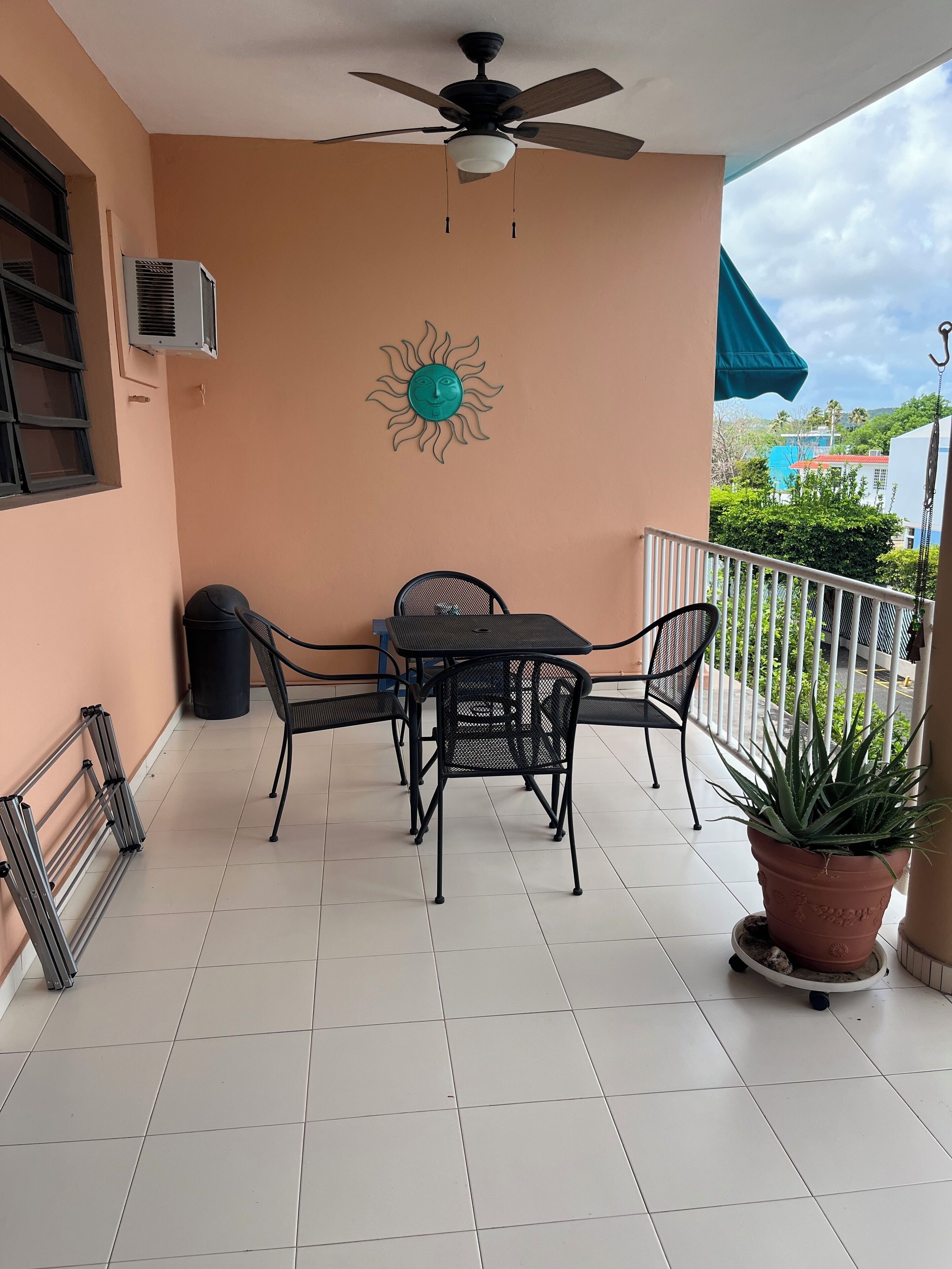 A balcony with a table and chairs, a ceiling fan, a wall-mounted sun decoration, and a potted plant.