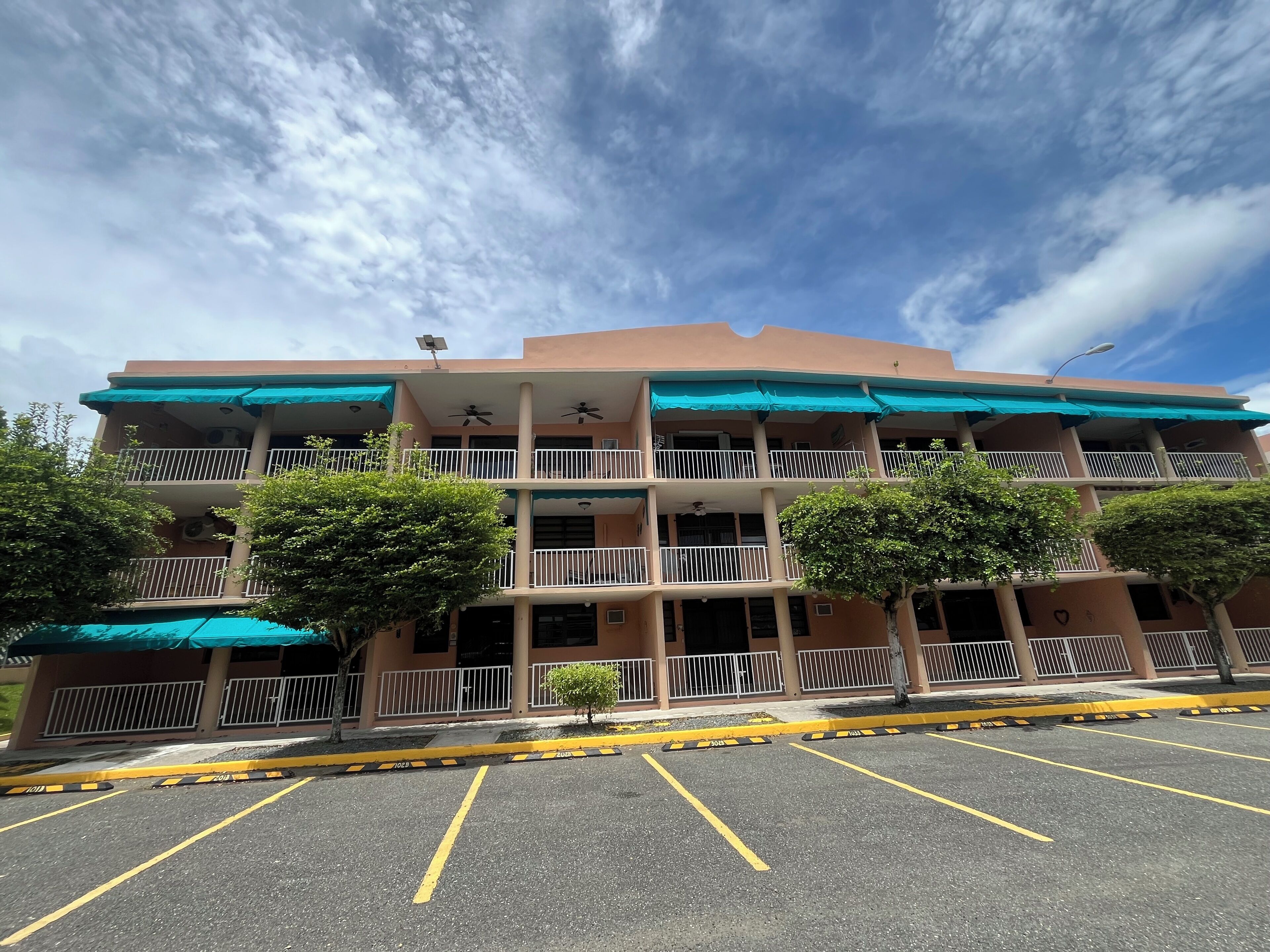 Un edificio de varios pisos con balcones, estacionamiento y árboles en la parte frontal.