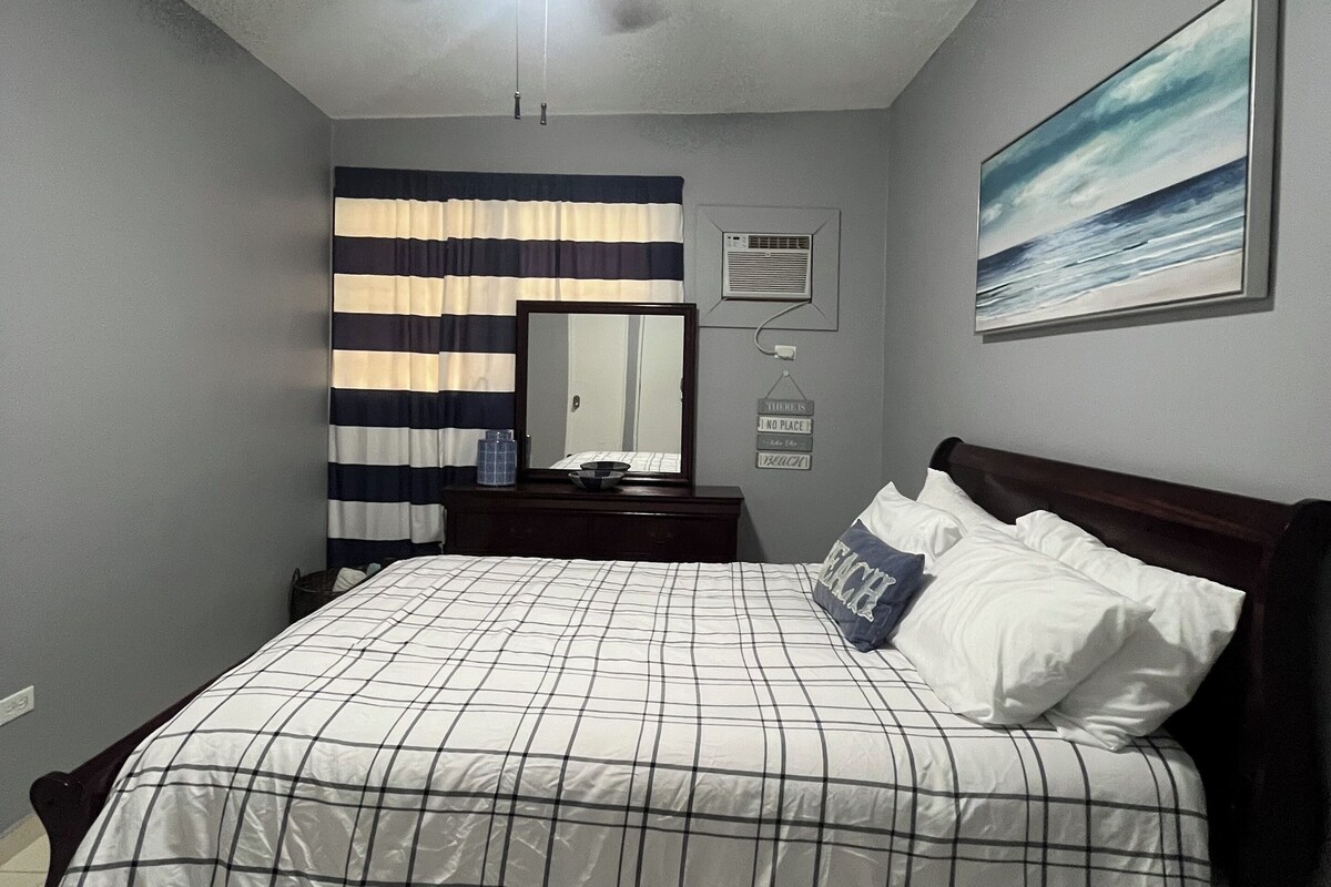 Quarto com cama, ventoinha no teto, janela com cortinas listradas, ar condicionado fixado na parede, quadro emoldurado e mesa de cabeceira com abajur.