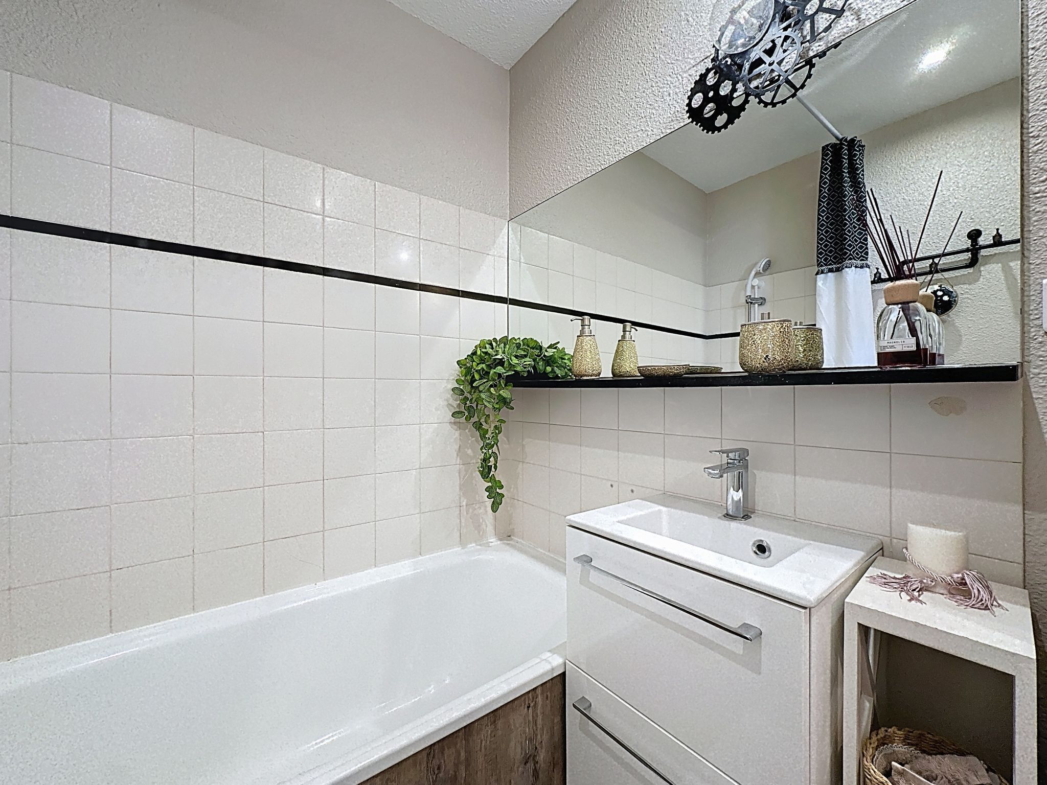 Een moderne badkamer met een witte badkuip, een wastafelmeubel met een wastafel en een spiegel erboven. Er is een plank met toiletartikelen en een hangplant.