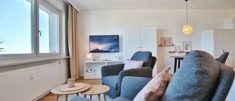 Uma sala de estar moderna com uma janela grande, uma TV de tela plana, um sofá, uma mesa de centro e uma escrivaninha com abajur.