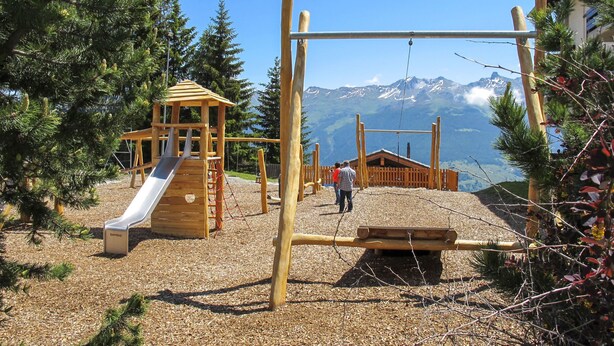 Un parque infantil con tobogán y columpios, rodeado de árboles y vistas a la montaña.