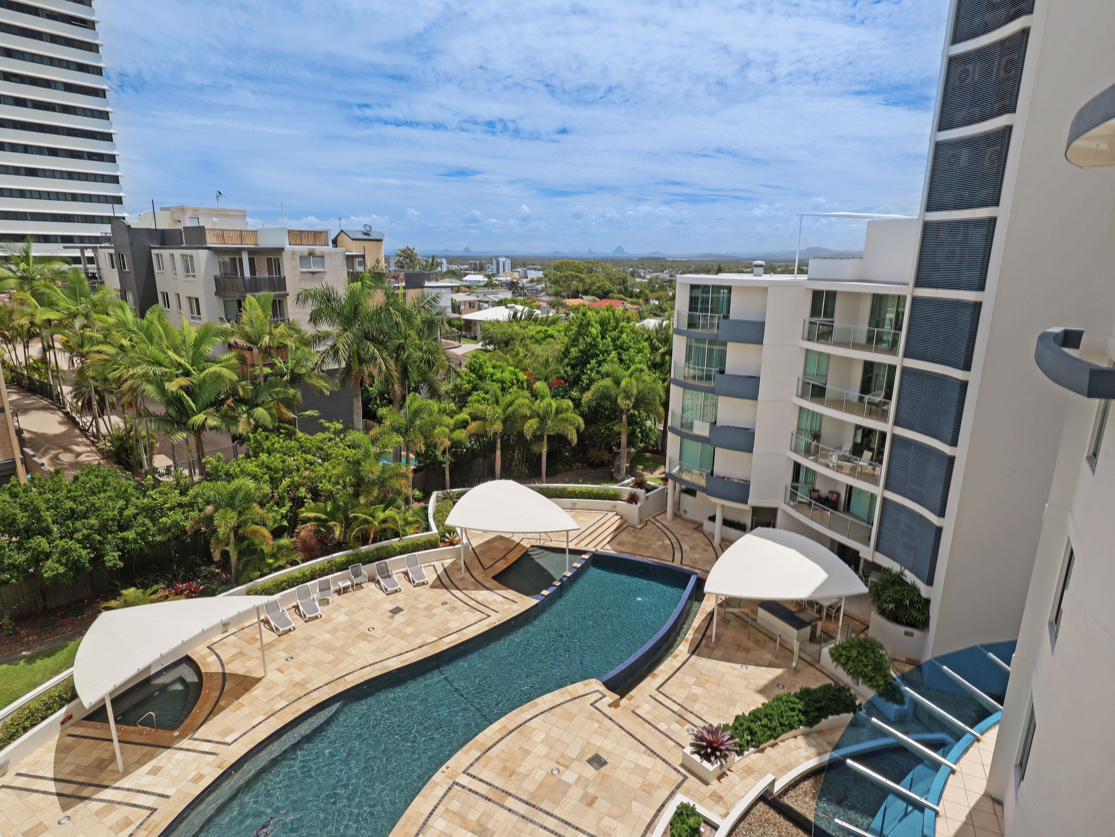 A rooftop pool area with lounge chairs and umbrellas, surrounded by greenery and modern buildings.