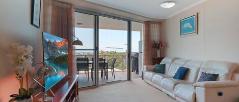 A living room with a sofa, a TV, a dining area with chairs, and a balcony with a view.