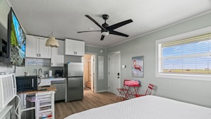 Een kleine hotelkamer met een bed, kitchenette, plafondventilator en een raam met zonwering.